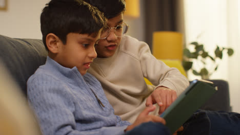 Two-Young-Boys-Sitting-On-Sofa-At-Home-Playing-Games-Or-Streaming-Onto-Digital-Tablet